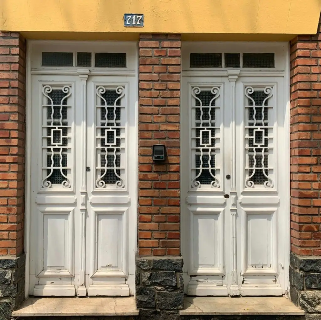 Une paire de portes doubles en bois blanc avec des ferronneries décoratives, mise en valeur par une restauration de briques qui allie architecture classique et savoir-faire artisanal.