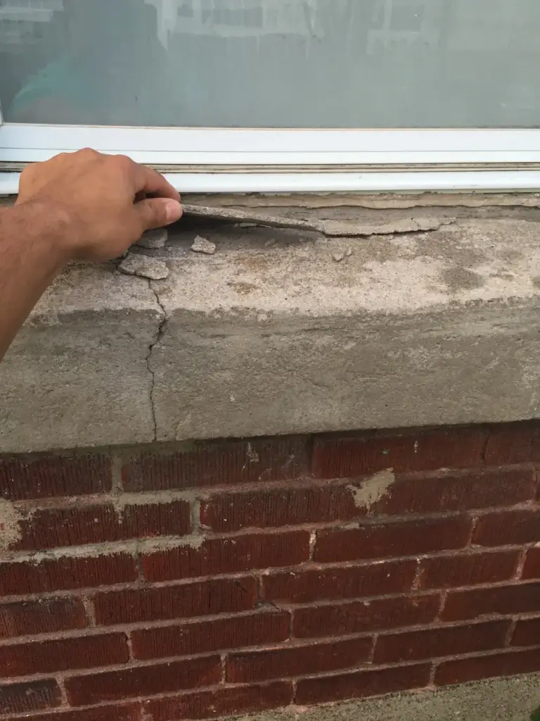 Gros plan sur une allège de fenêtre en béton fissurée et friable, avec une personne soulevant une section détachée, montrant des signes clairs de détérioration.